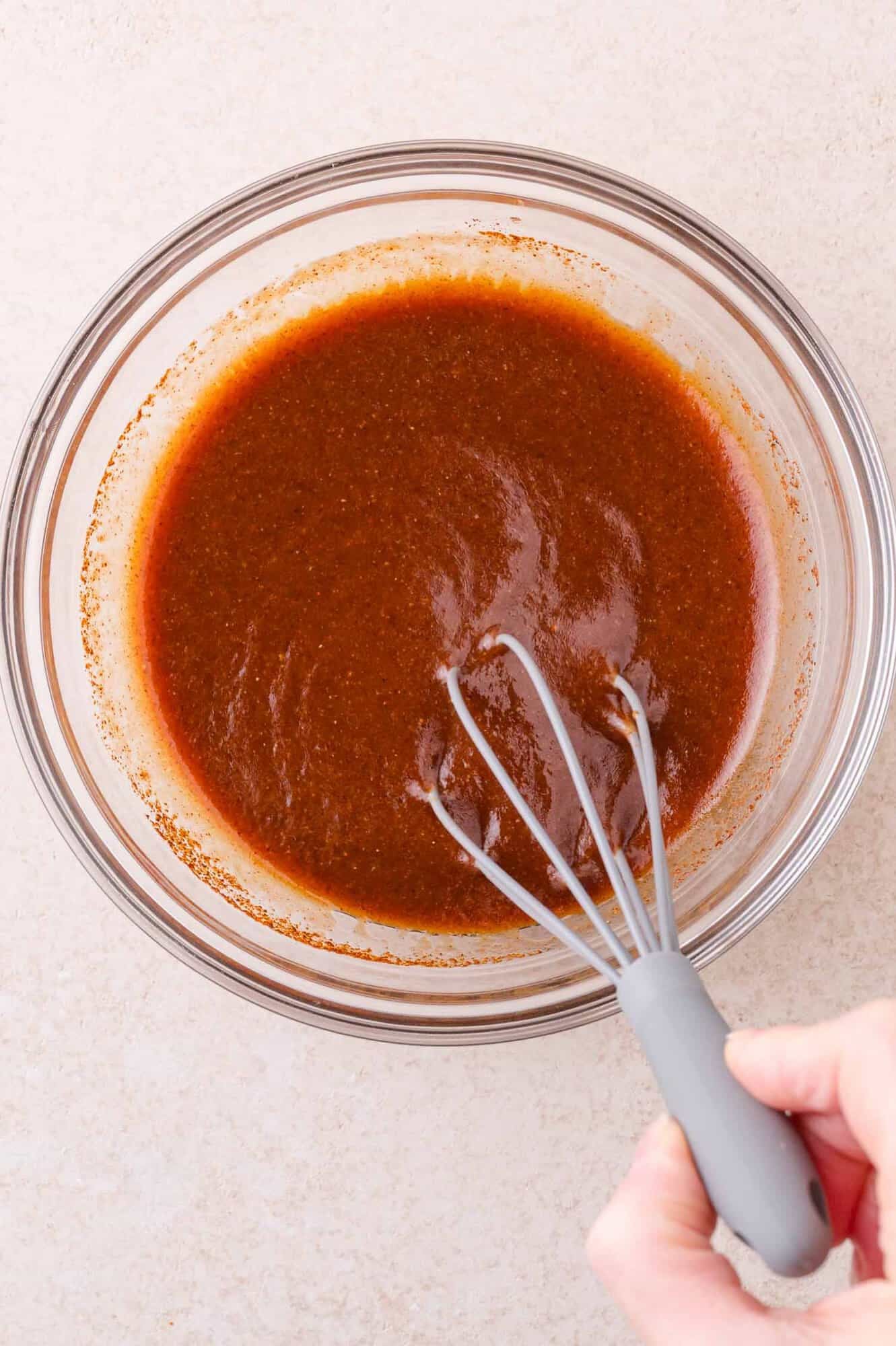 A hand holding a whisk mixes barbecue marinade in a clear glass bowl on a light countertop.
