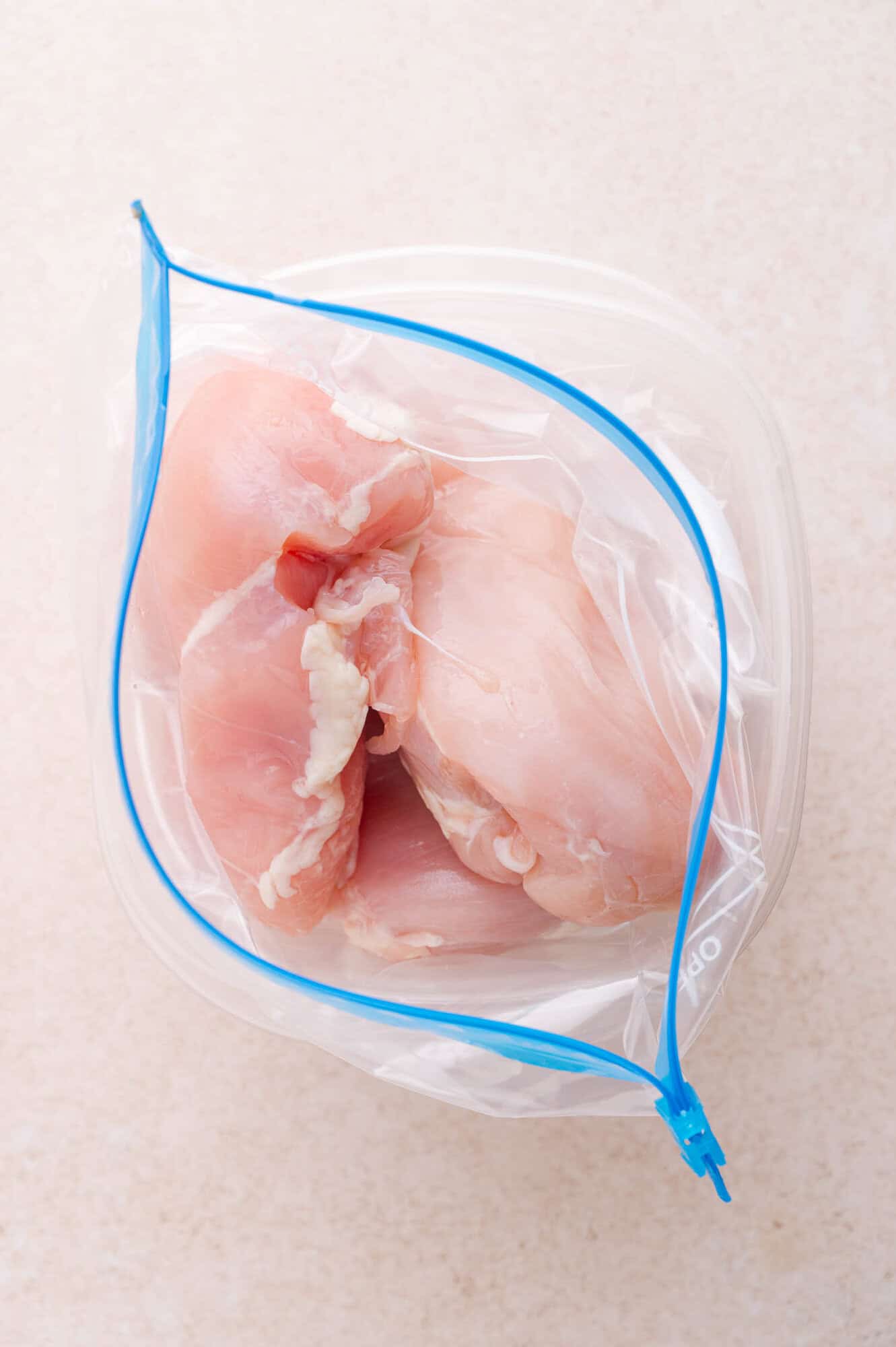 Raw chicken breasts in a clear resealable plastic bag on a light-colored surface.