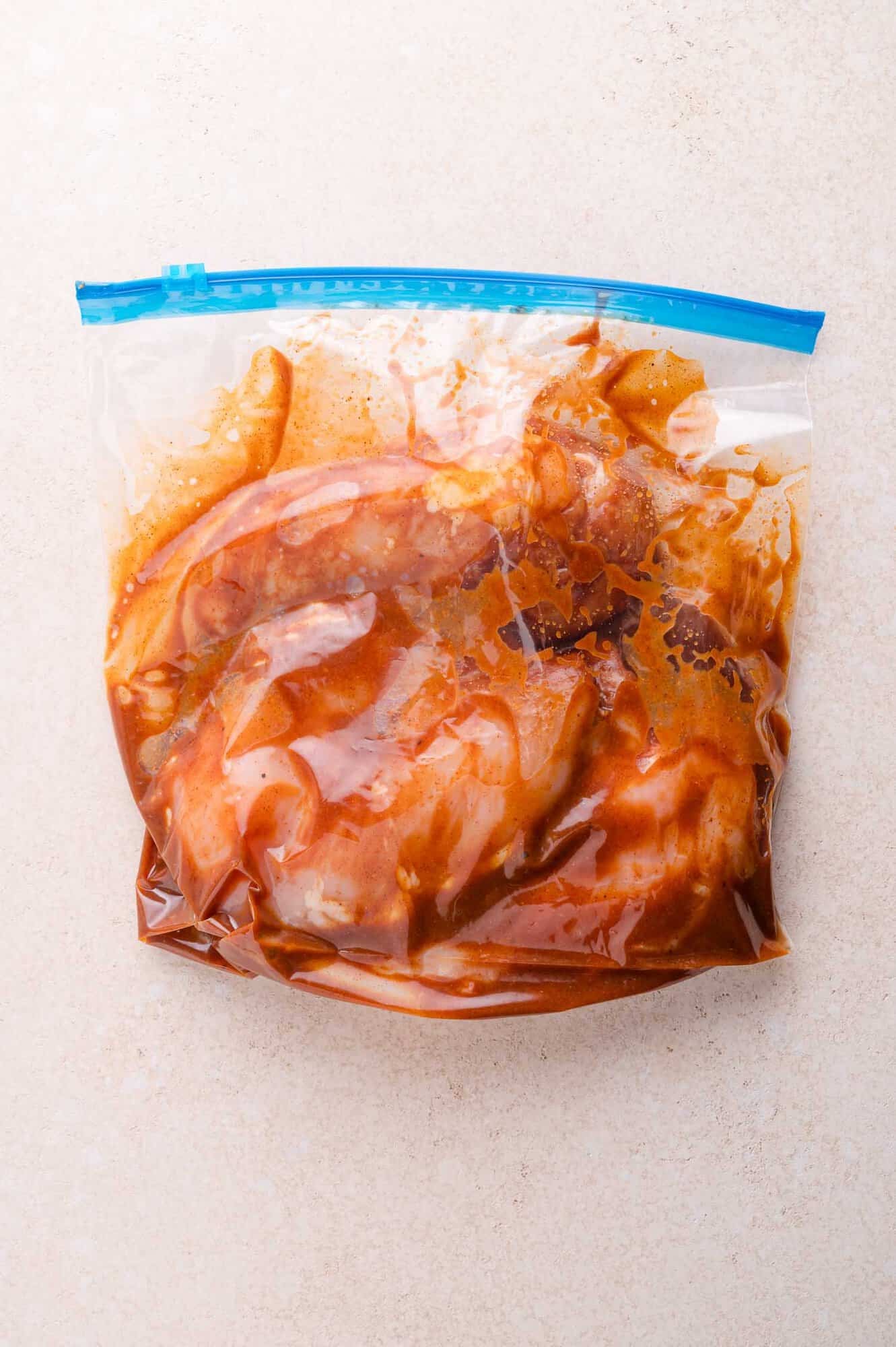 A resealable plastic bag containing raw meat marinating in a bbq marinade on a light-colored surface.