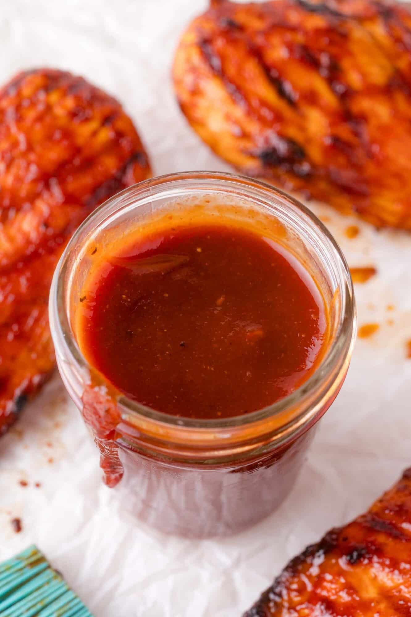 A glass jar filled with barbecue sauce sits on a white surface, surrounded by pieces of grilled chicken.