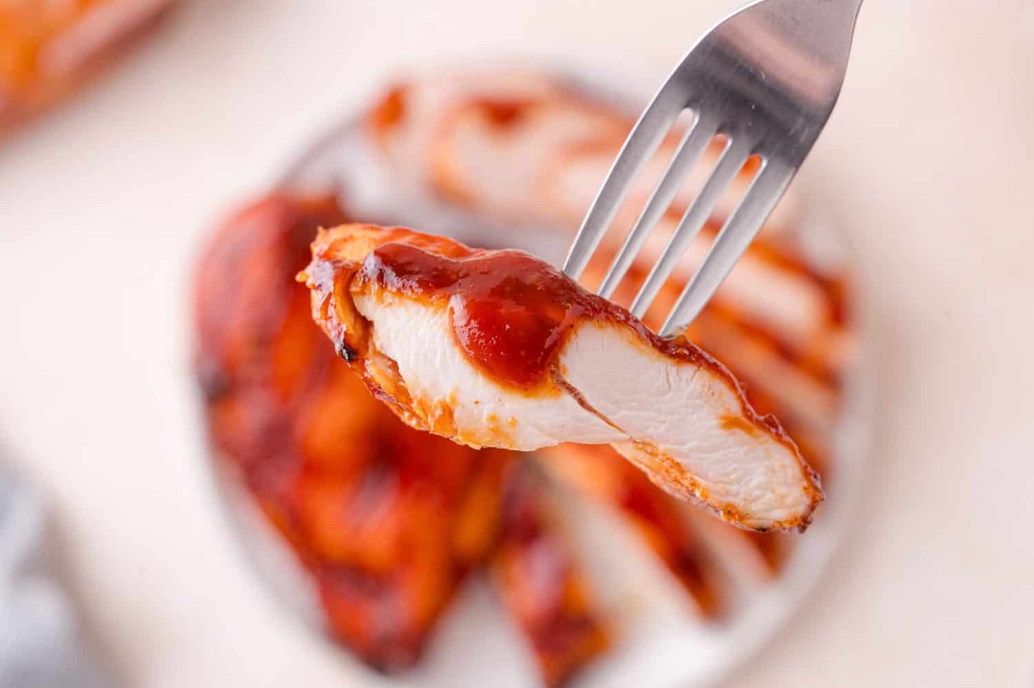 A fork holds a saucy slice of cooked chicken breast above a plate with more sliced chicken in the background.
