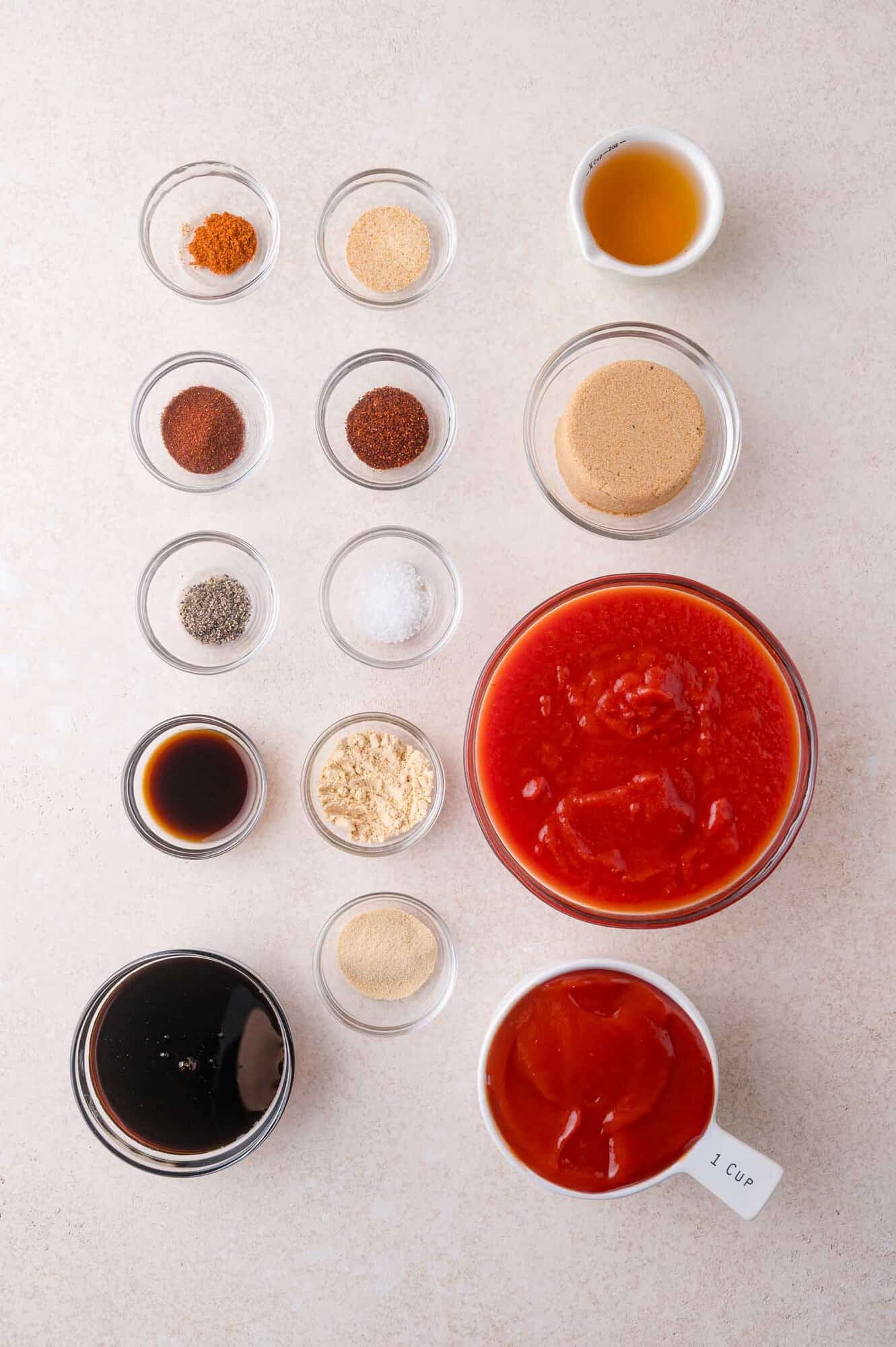 Ingredients for homemade barbecue sauce arranged on a white surface, including spices, brown sugar, vinegar, Worcestershire sauce, molasses, tomato sauce, and crushed tomatoes.
