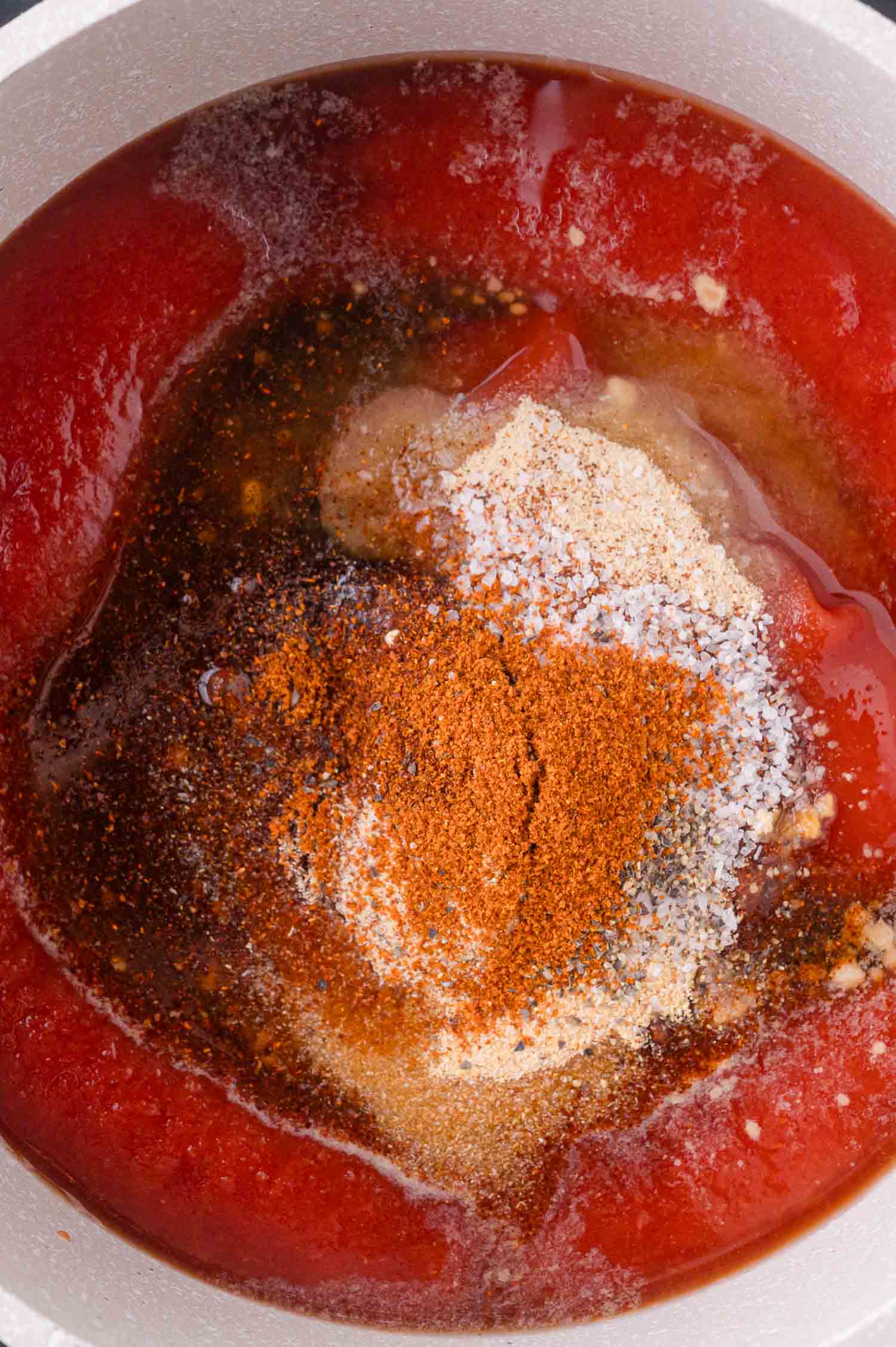 A close-up of a pot containing barbeque sauce with various spices and seasonings sprinkled on top, ready to be mixed.