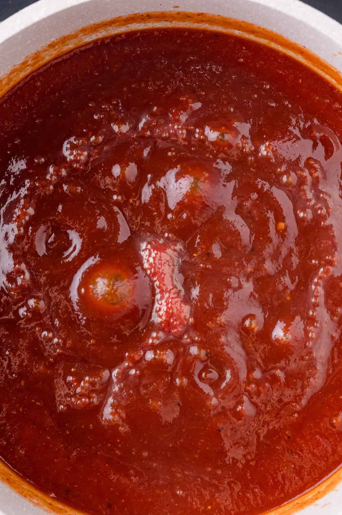 A close-up view of barbecue sauce simmering in a pot, with visible whole tomatoes and a rich, red texture.