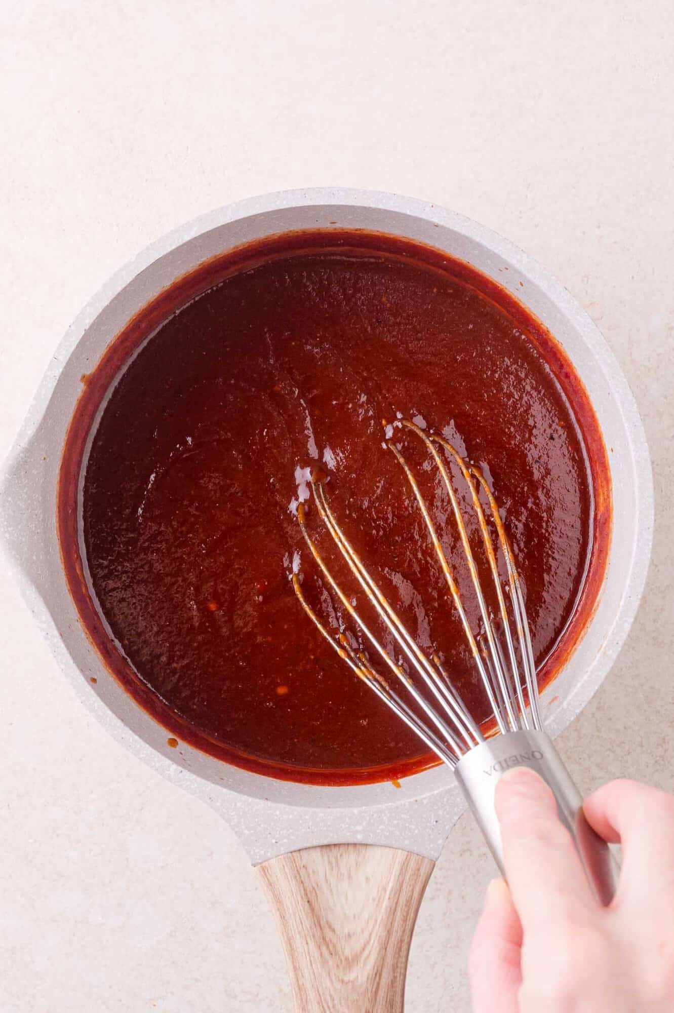 A hand uses a whisk to stir a pot of thick, dark red BBQ sauce on a light countertop.