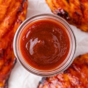 A glass jar filled with barbecue sauce is centered, surrounded by pieces of grilled chicken on a white surface.