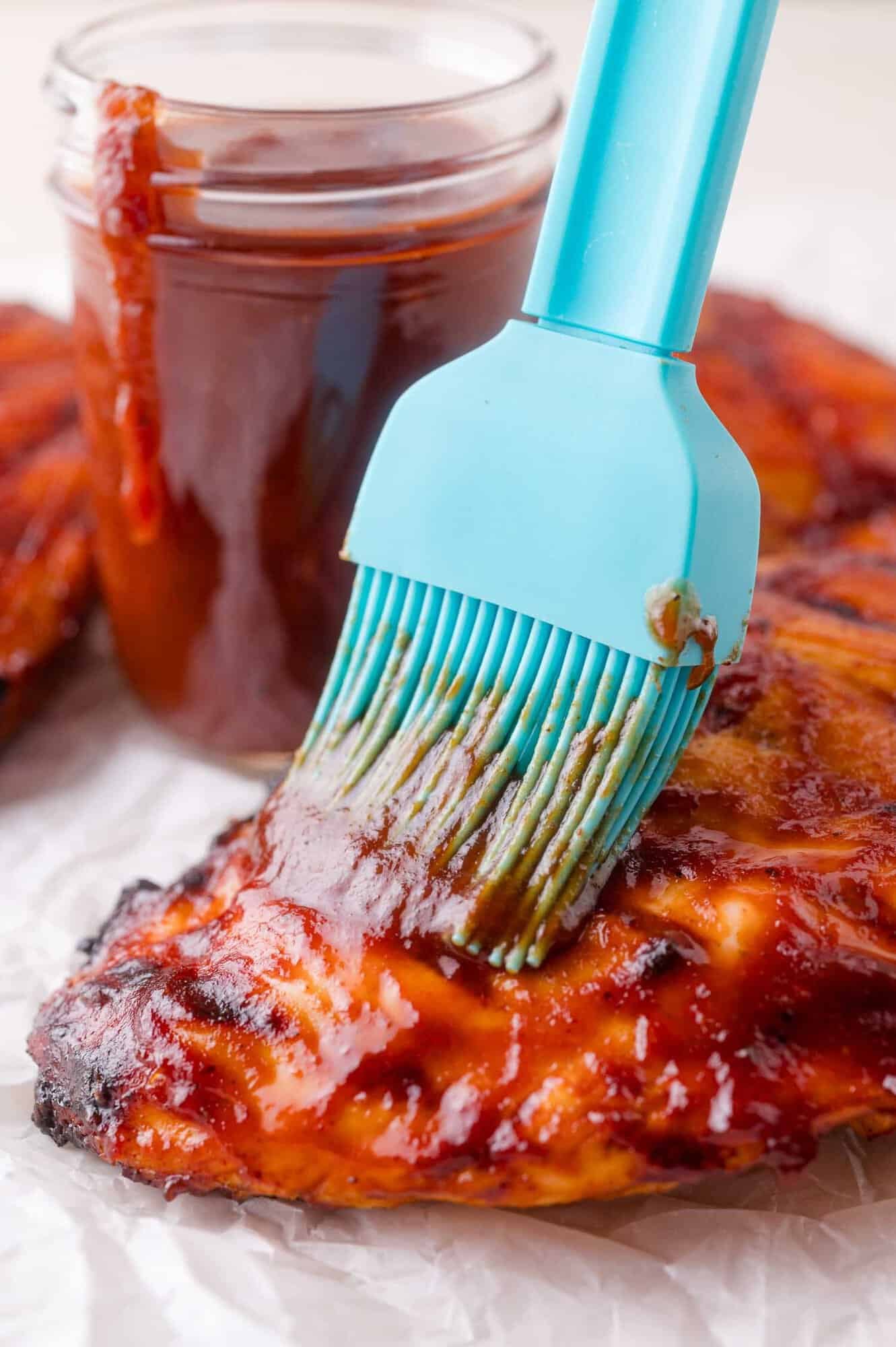 A blue silicone brush applies barbecue sauce onto grilled chicken, with a jar of barbecue sauce in the background.
