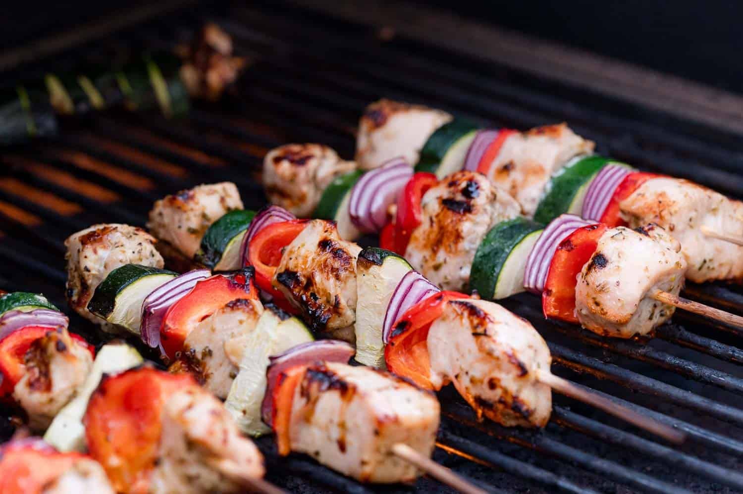 Chicken and vegetable skewers, including zucchini, red onion, and red bell pepper, cooking on a grill.