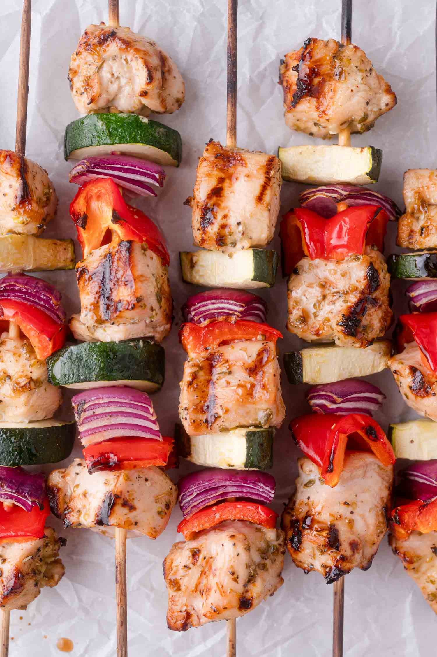 Grilled chicken kebabs with pieces of zucchini, red bell pepper, and red onion on wooden skewers, arranged on a white surface.