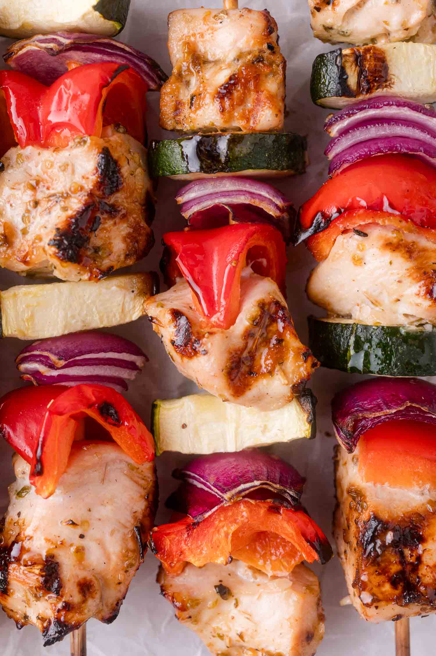 Grilled chicken skewers with pieces of red bell pepper, zucchini, and red onion on wooden sticks, arranged closely together.