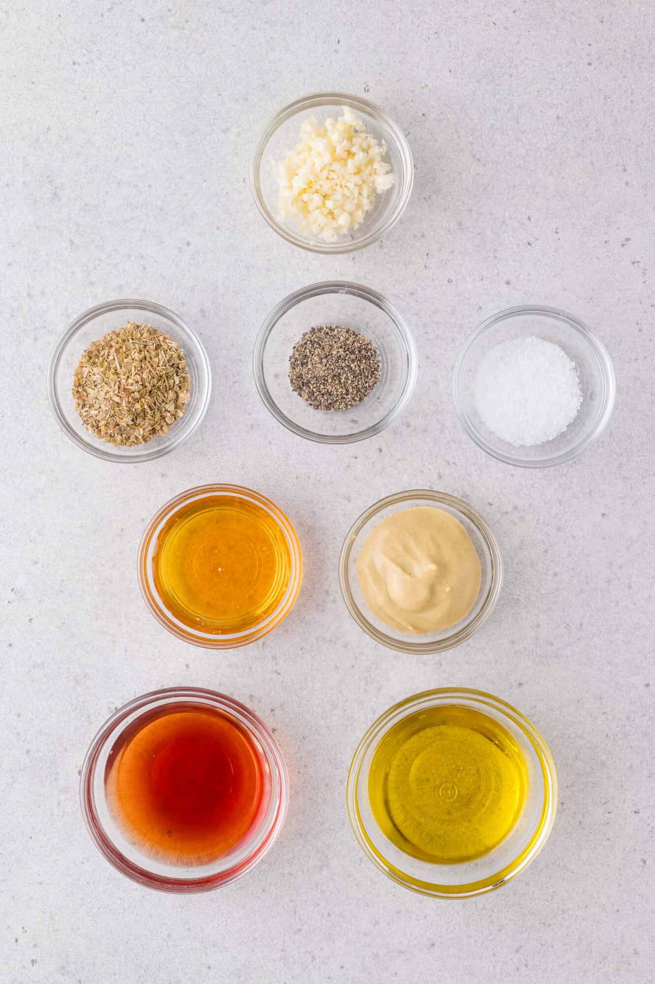 Nine small glass bowls containing minced garlic, dried herbs, black pepper, salt, honey, Dijon mustard, vinegar, and olive oil are arranged on a light surface.