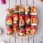 Four grilled chicken and vegetable kabobs with pieces of chicken, red onion, zucchini, and red bell pepper arranged on a round plate.