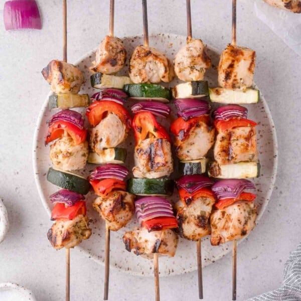 Four grilled chicken and vegetable kabobs with pieces of chicken, red onion, zucchini, and red bell pepper arranged on a round plate.