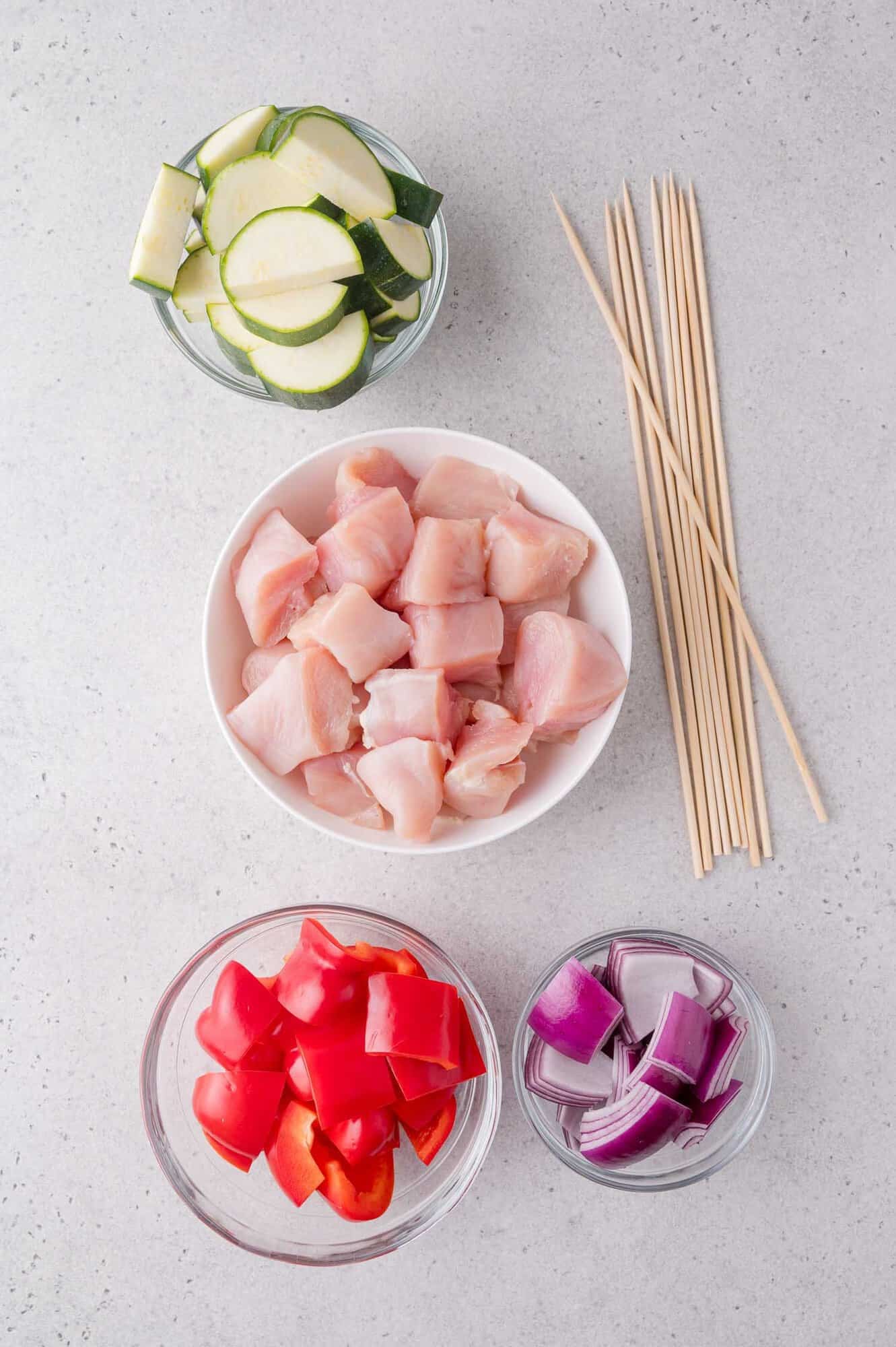 Bowls of chopped zucchini, raw chicken, red bell pepper, and red onion with wooden skewers arranged on a light surface.