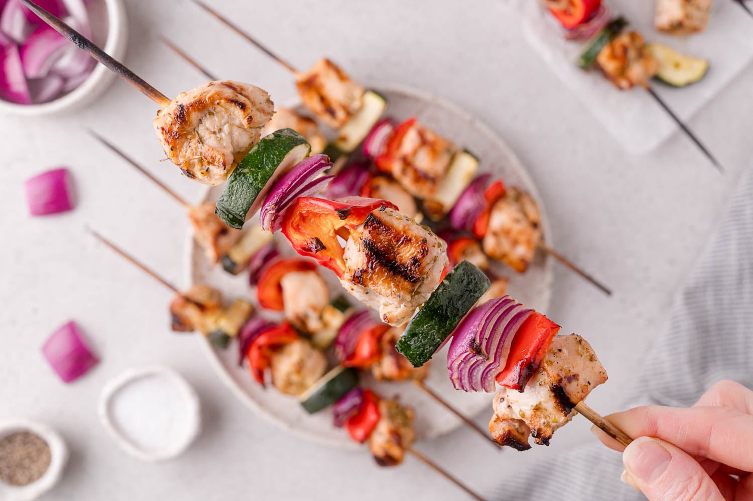 A hand holds a grilled skewer with pieces of chicken, zucchini, red onion, and red bell pepper over a plate of similar skewers on a light surface.