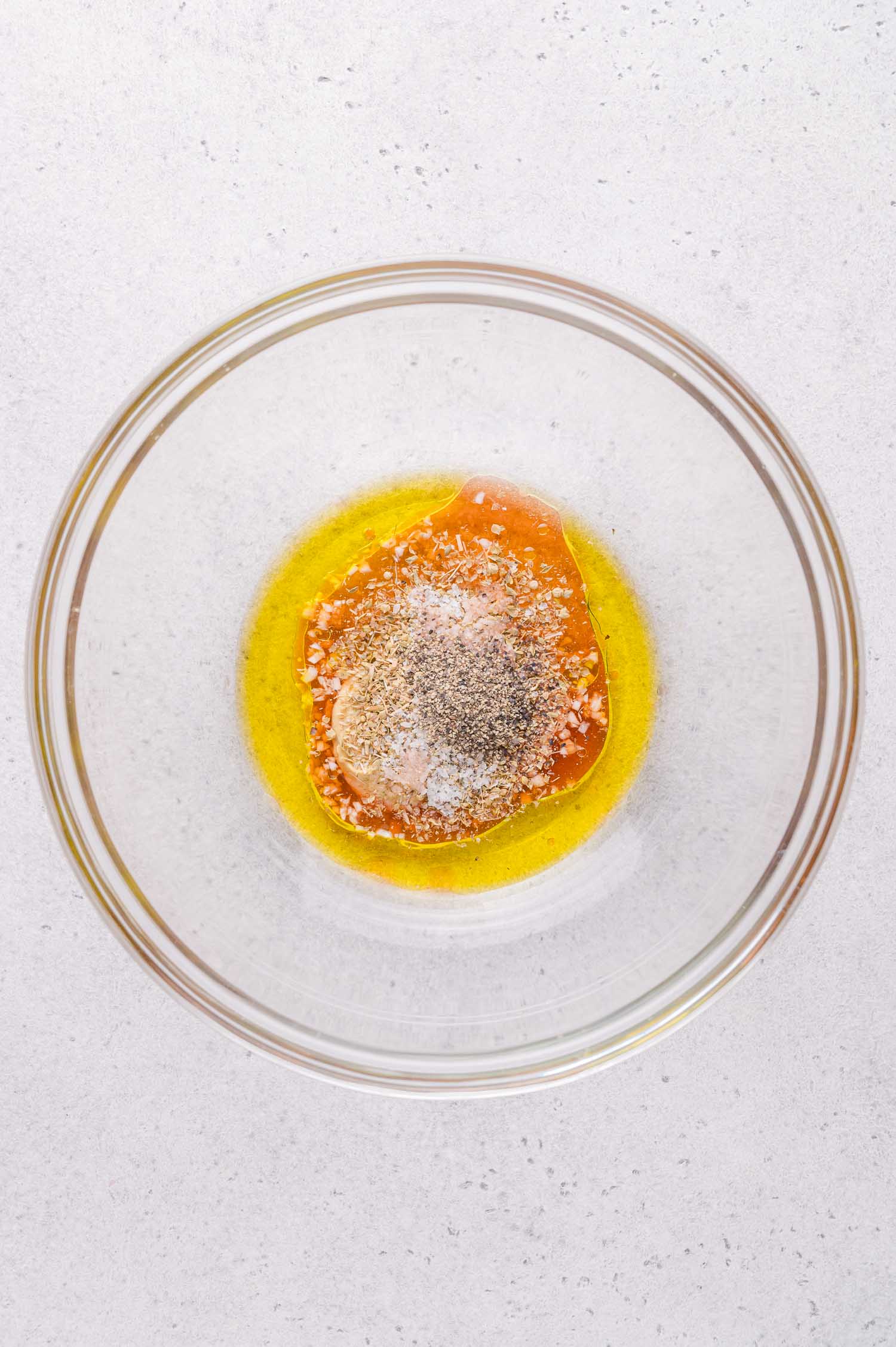 A clear glass bowl containing olive oil, honey, and seasonings on a light countertop.