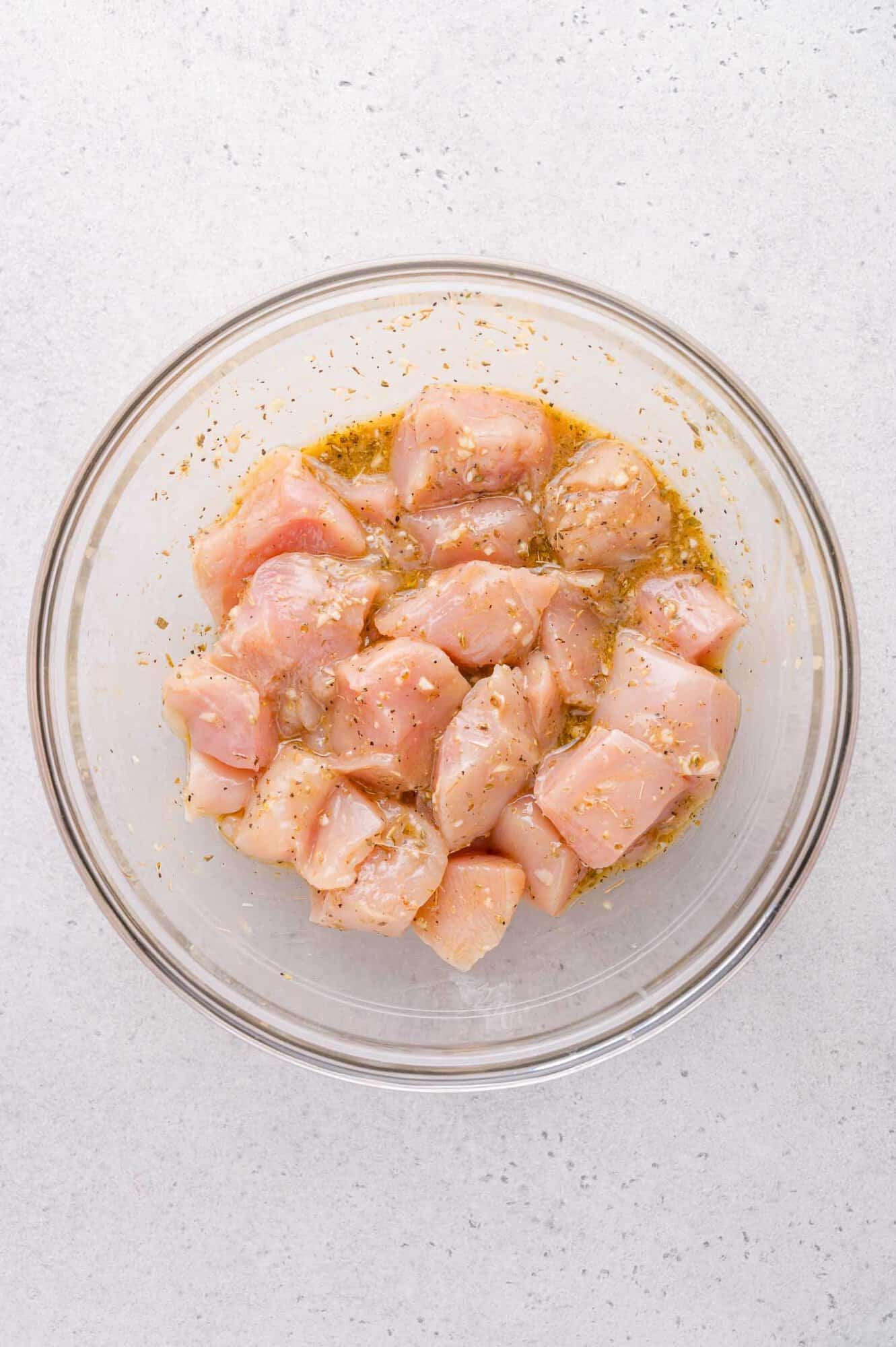 A glass bowl filled with raw, cubed chicken marinating in oil and seasonings on a light-colored surface.