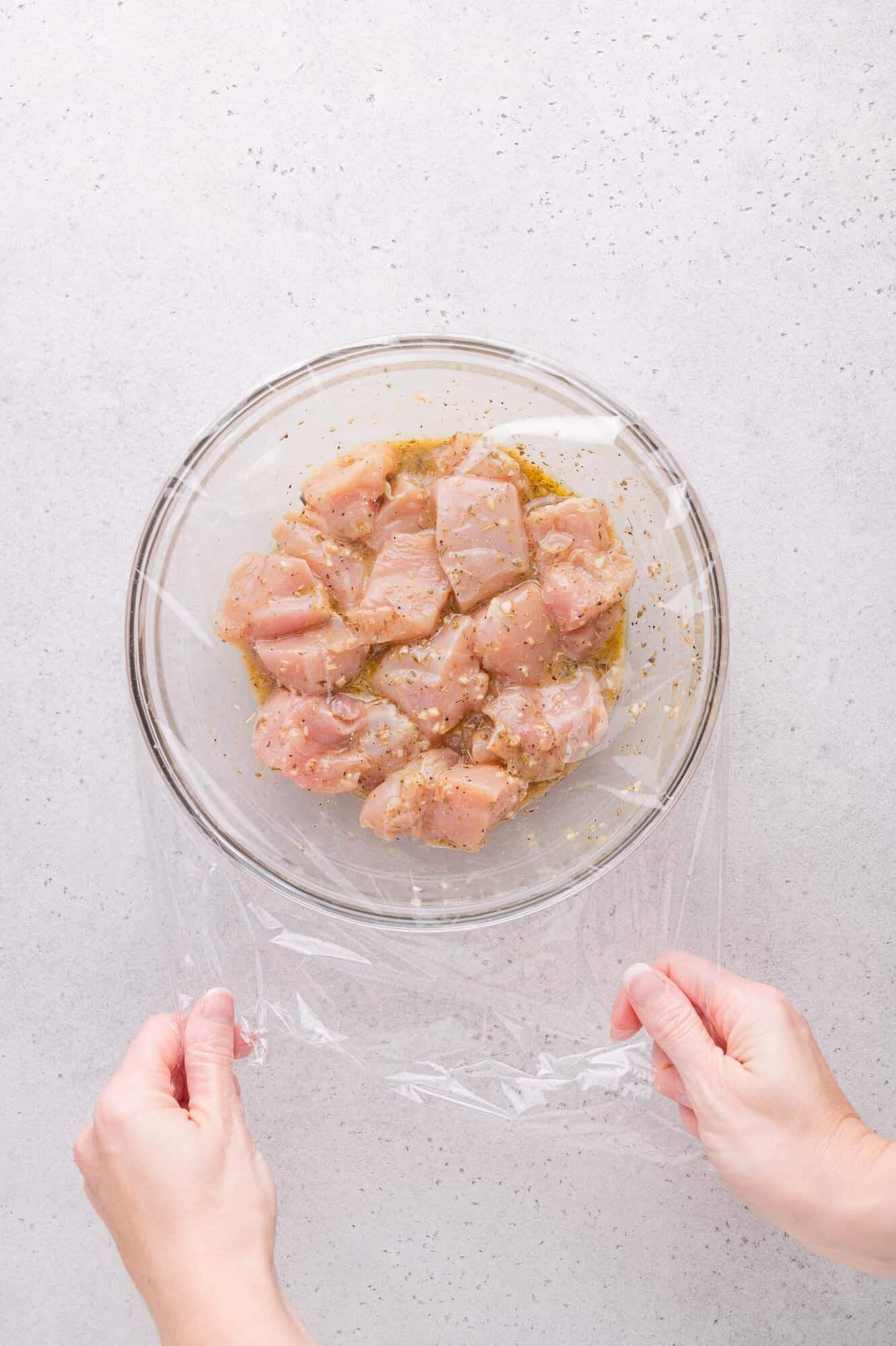 A pair of hands covers a glass bowl of marinated raw chicken pieces with plastic wrap on a light gray surface.