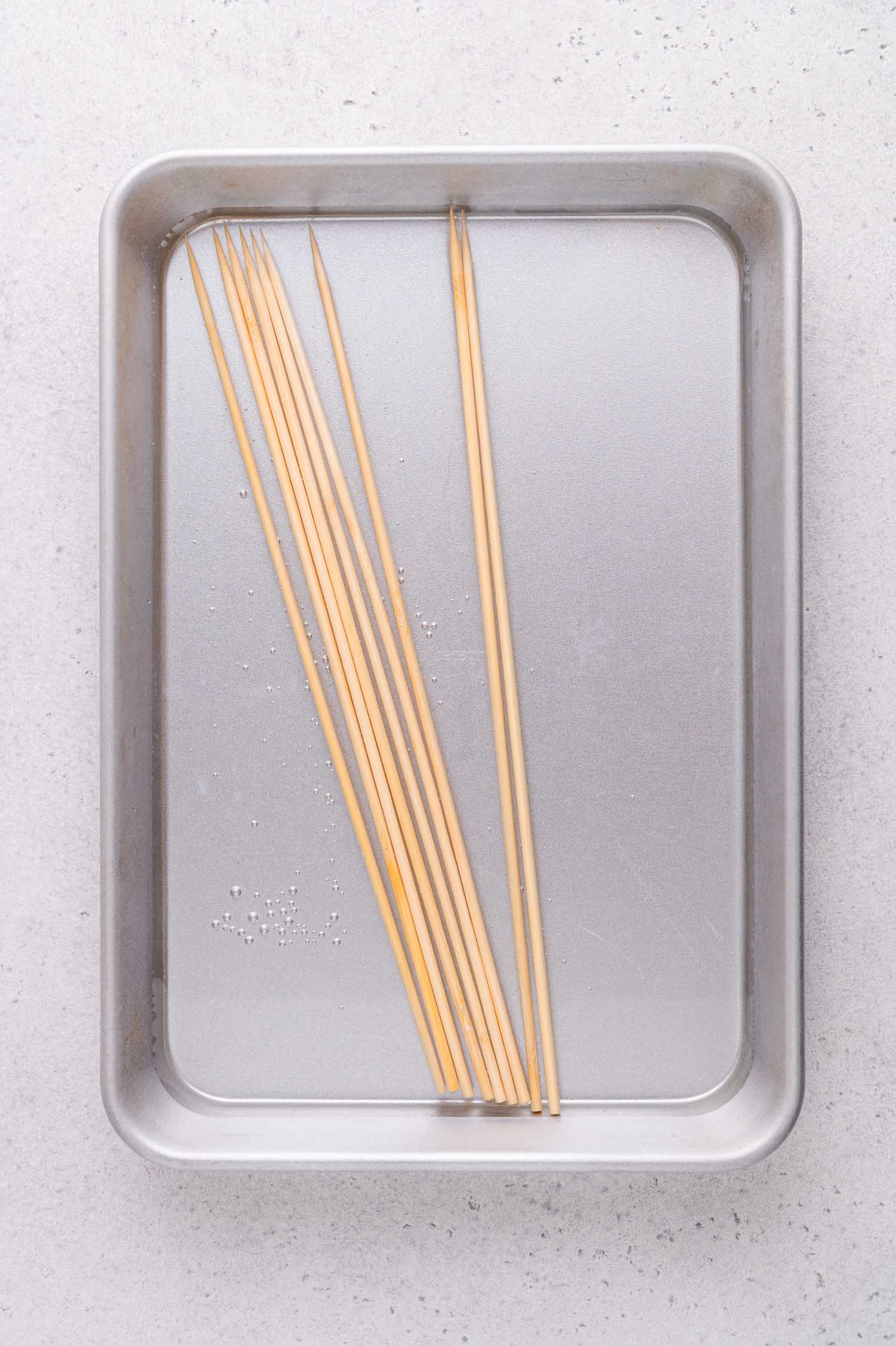 Wooden skewers soaking in water in a metal baking tray on a light-colored surface.