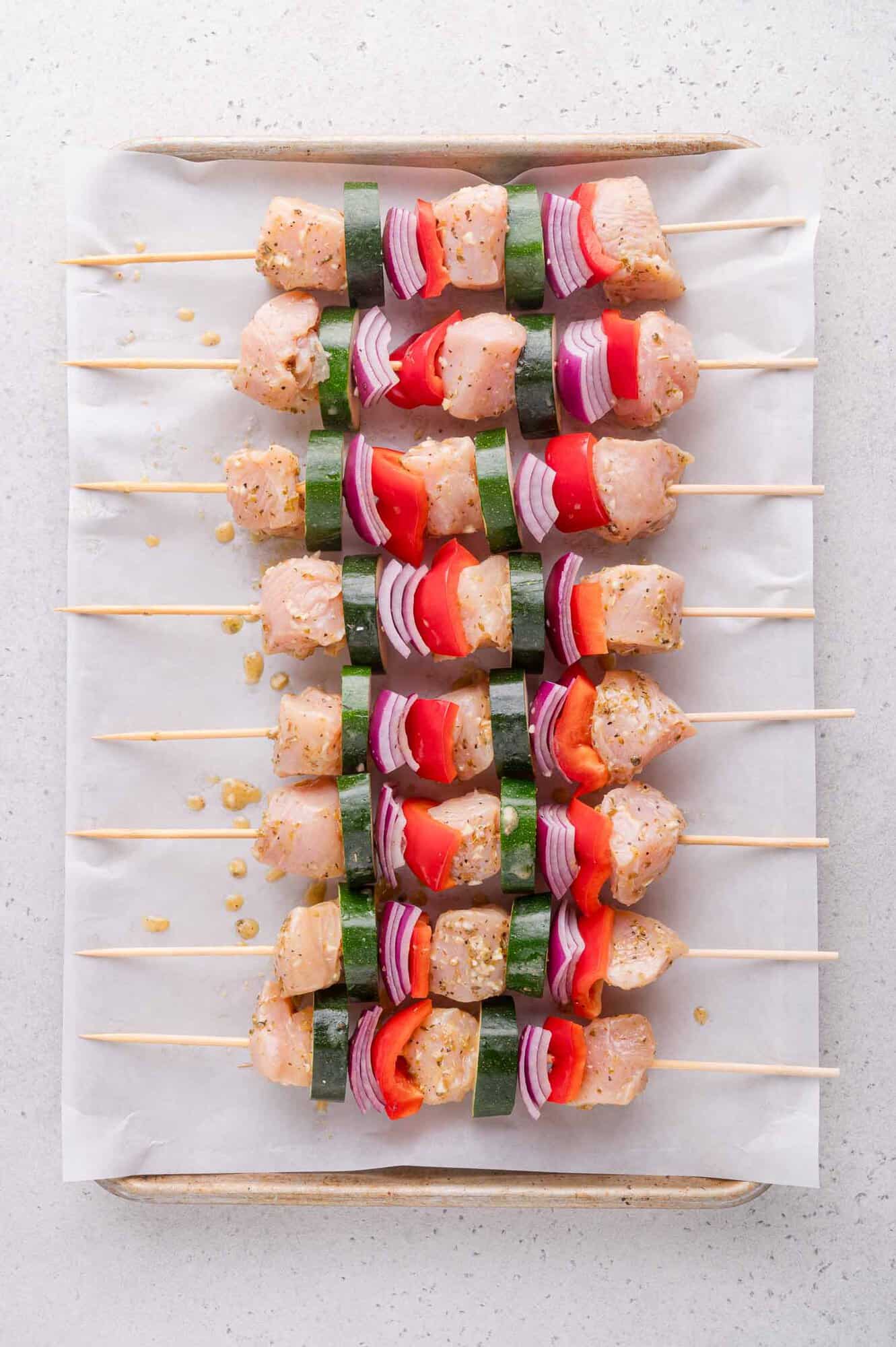 Raw skewers with chunks of chicken, zucchini, red onion, and red bell pepper arranged neatly on a parchment-lined baking sheet.