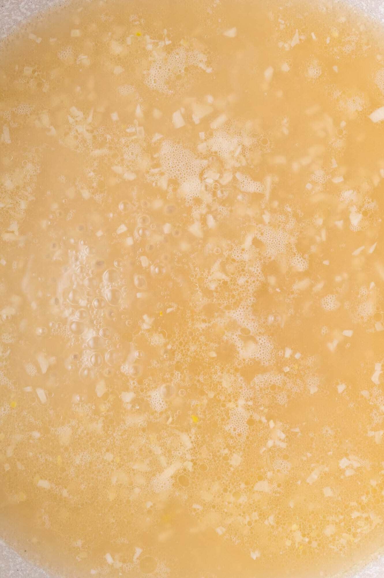 Close-up of a light yellow liquid with small chopped garlic pieces and bubbles floating on the surface.