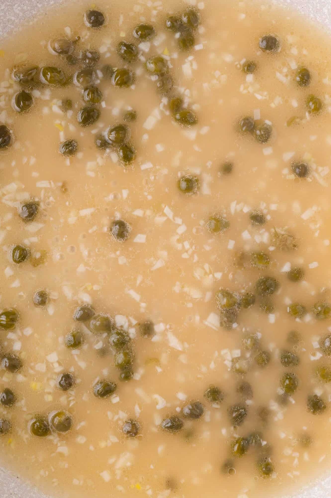 Close-up of a light brown sauce with capers and finely chopped garlic mixed throughout.