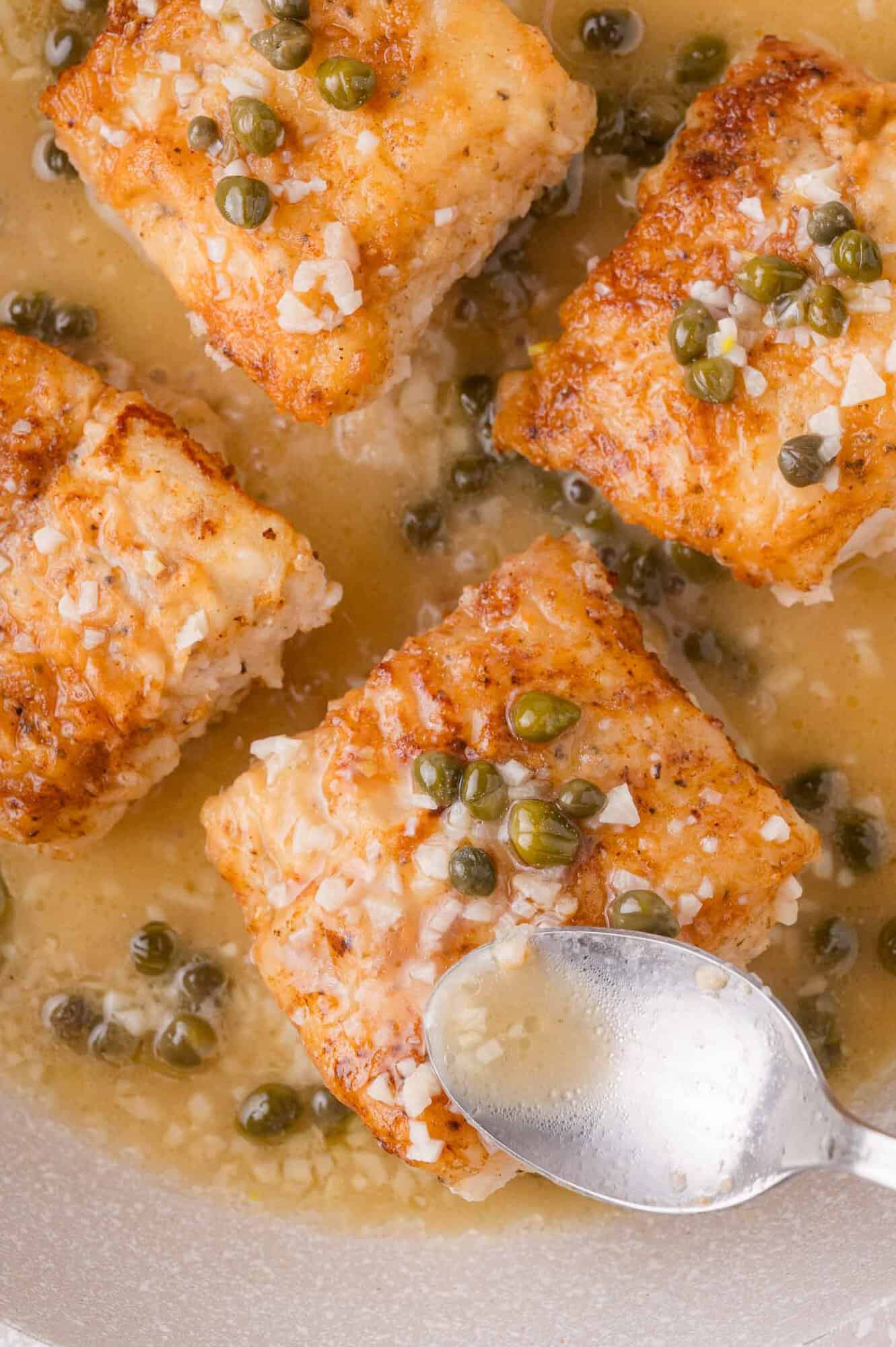 Four pieces of cooked fish in a lemon butter sauce, topped with capers and coarse salt, with a spoon drizzling sauce over one piece.