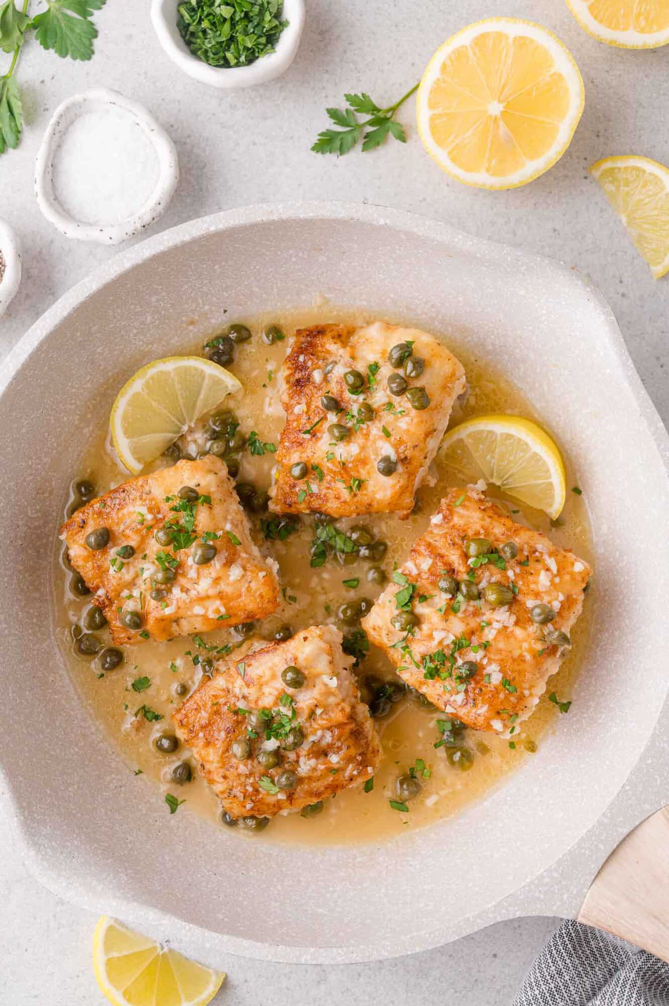 Four pieces of pan-seared fish in a light sauce with capers and lemon slices, garnished with chopped parsley, in a beige skillet on a light countertop.