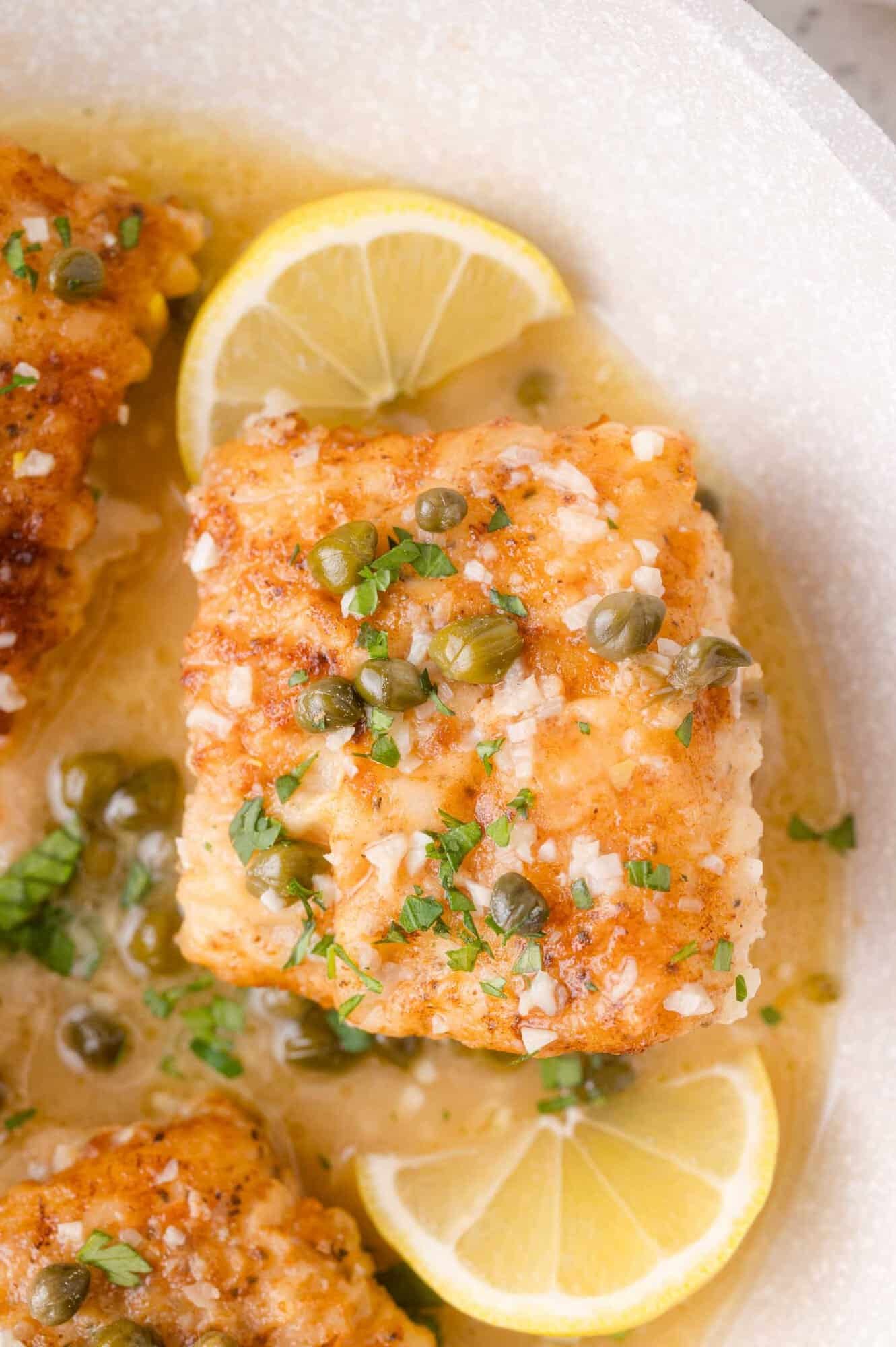 A piece of cooked cod topped with capers and herbs, served in a light sauce with lemon wedges on a white plate.