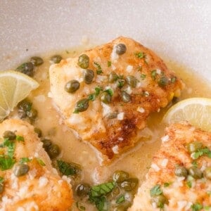 Pan-seared cod fillet topped with capers, chopped herbs, and a lemon butter sauce, garnished with lemon slices.
