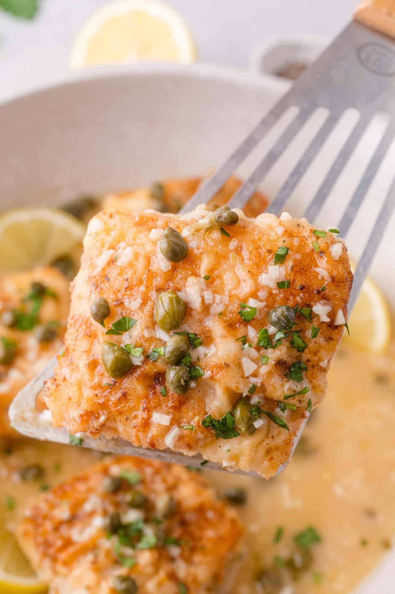 A spatula lifts a piece of cooked cod piccata topped with capers, parsley, and a lemon butter sauce, with lemon slices visible in the background.