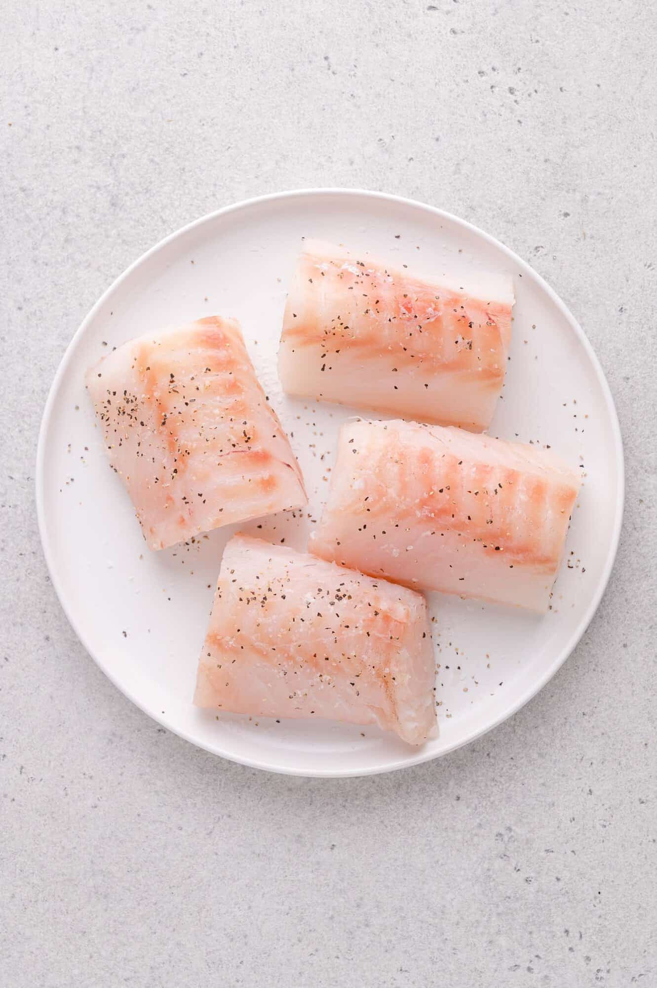 Four raw cod fillets, lightly seasoned with black pepper, are arranged on a white plate set on a light gray surface.