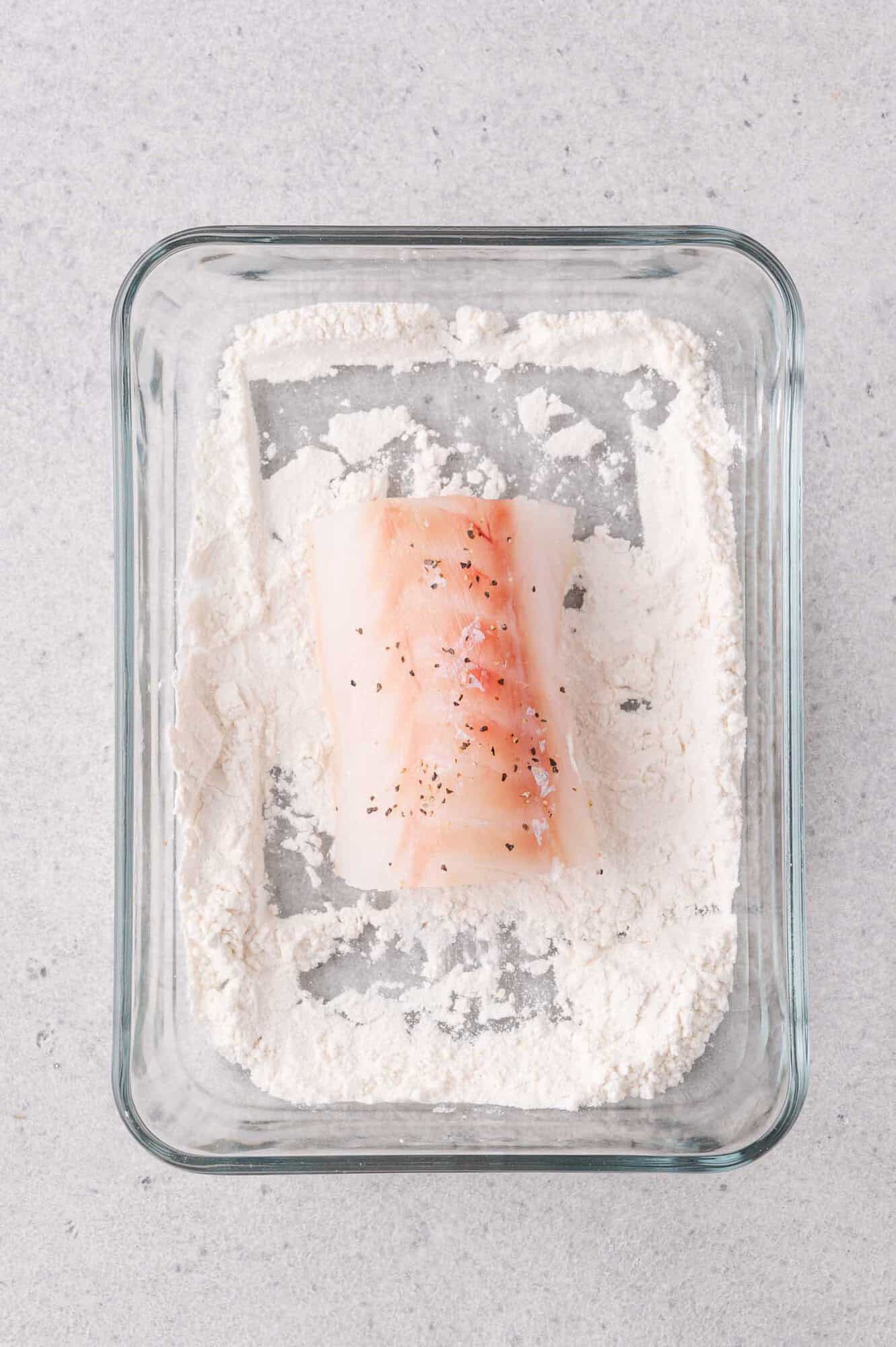 A raw fillet of cod seasoned with pepper sits in a glass dish filled with flour on a light gray surface.