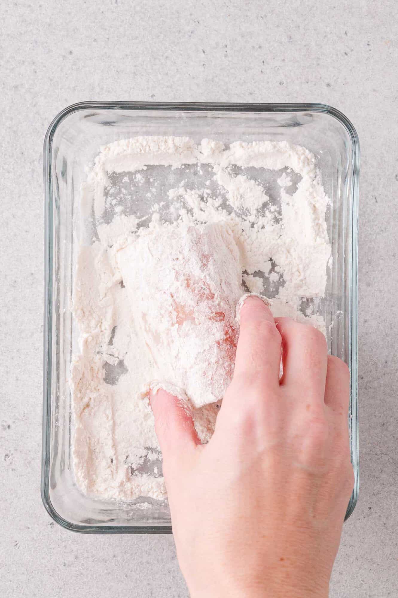 A hand coats a piece of raw cod with flour in a glass dish on a light-colored surface.