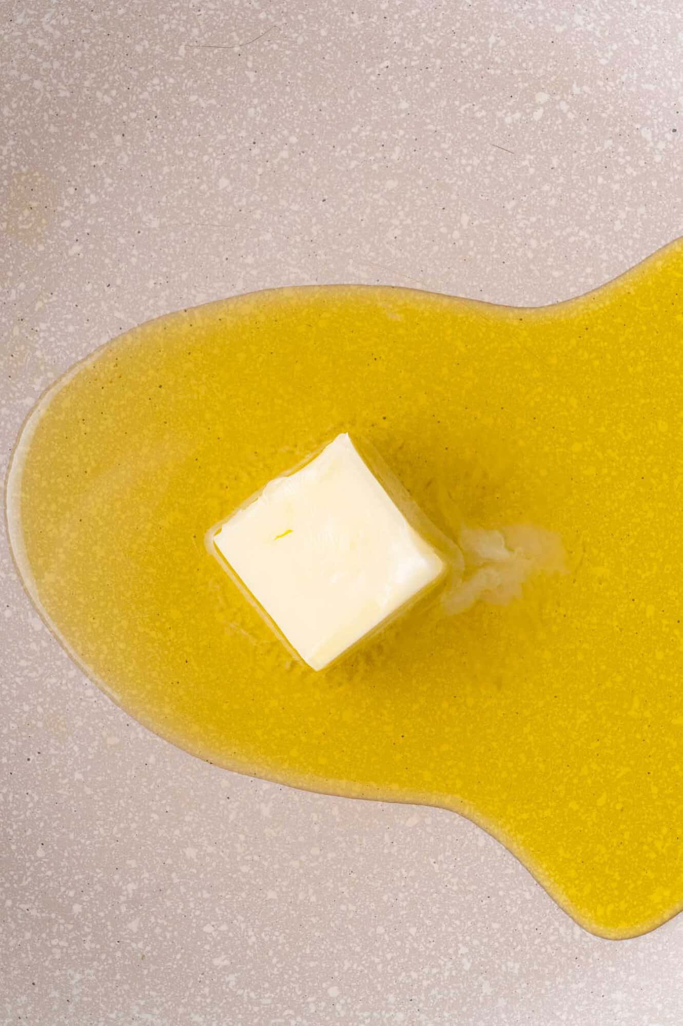 A cube of butter melting on a light-colored, speckled surface.