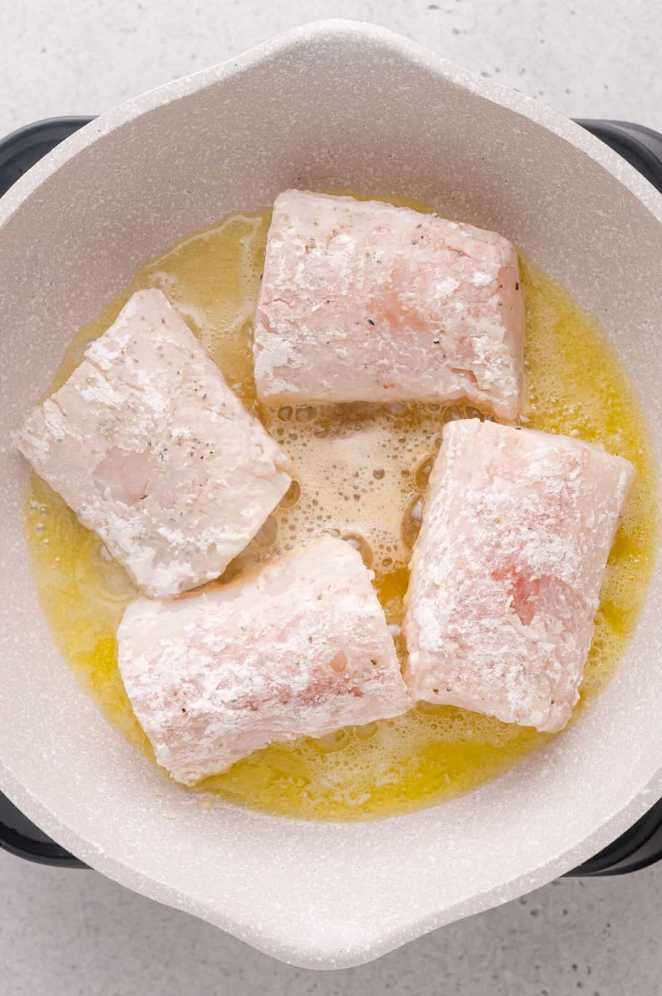 Four pieces of floured cod are being cooked in a light-colored pan with melted butter.