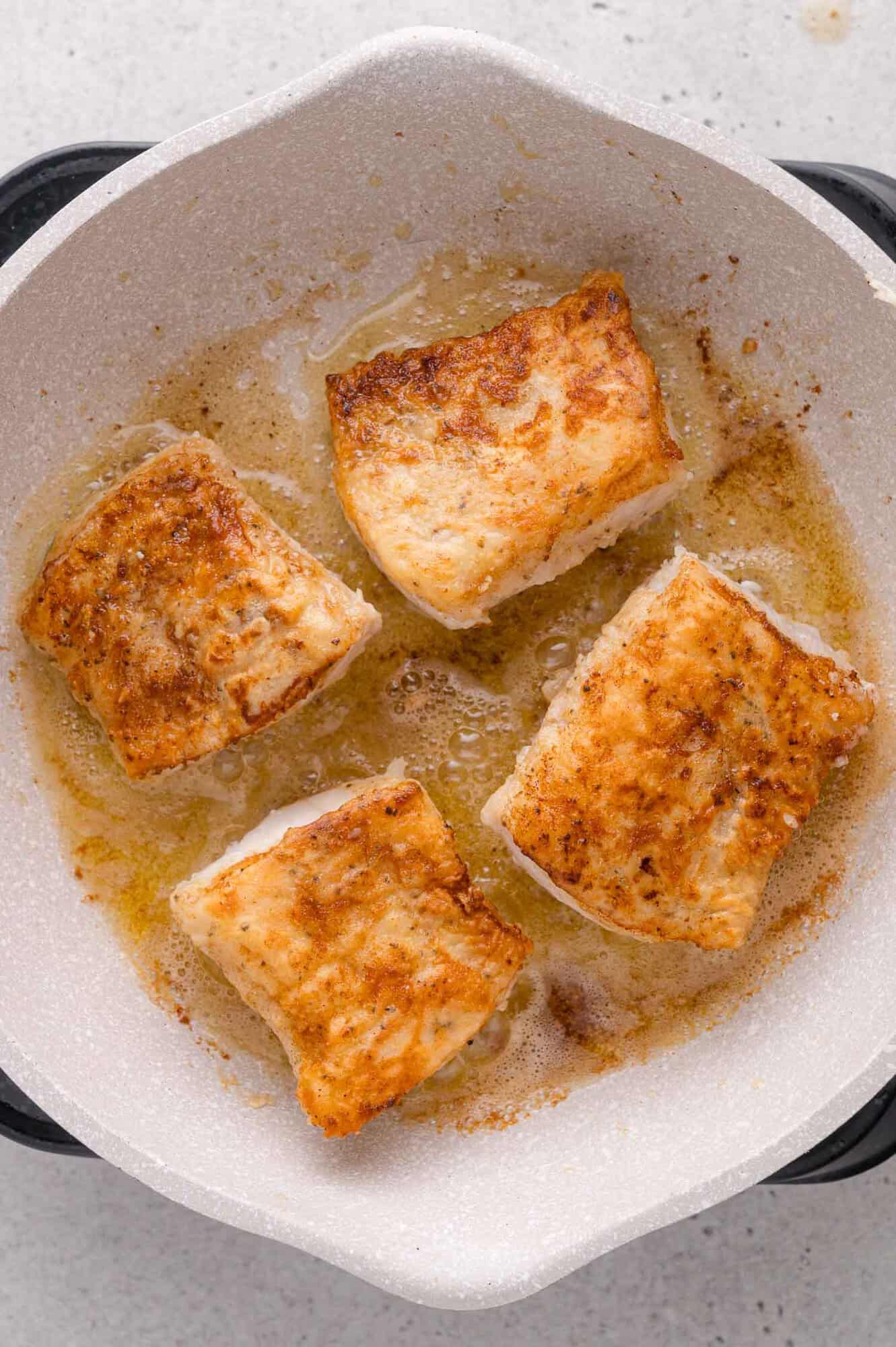 Four pieces of seasoned cod are being pan-seared in a white skillet with oil and browned edges.