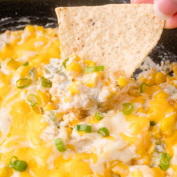 A hand dips a tortilla chip into a cheesy corn and jalapeño dip topped with sliced green onions.