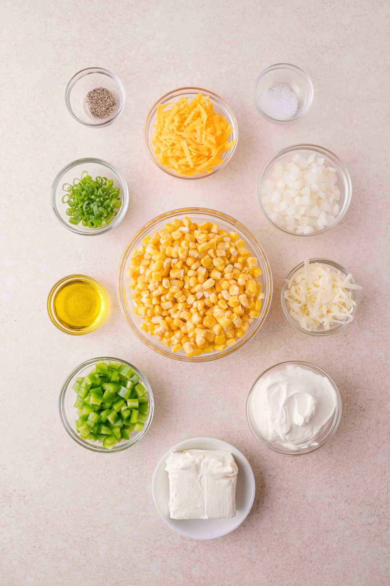 Overhead view of bowls containing corn, shredded cheese, diced onion, chopped jalapeno pepper, green onions, cream cheese, sour cream, oil, salt, and pepper on a light surface.