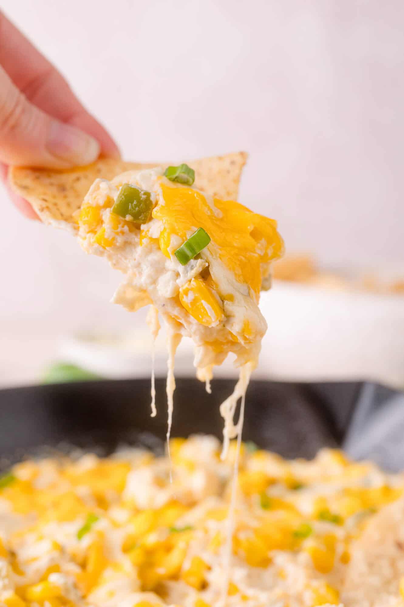 A hand holds a tortilla chip dipped in a cheesy, yellow and green onion-topped dip, with melted cheese stretching from the dish below.