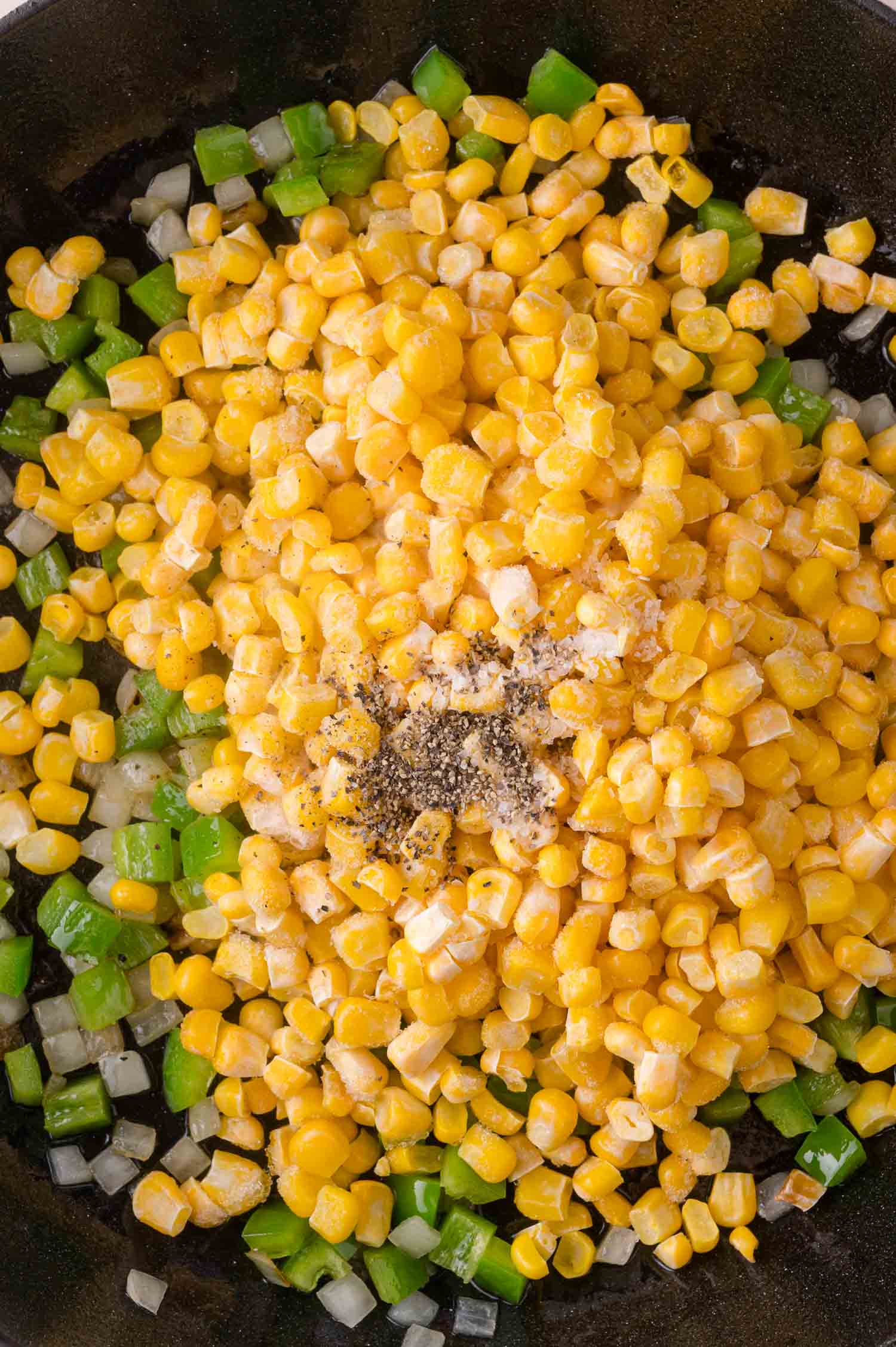 Corn, diced pepper, and chopped onion cooking in a skillet, topped with ground black pepper and seasoning.