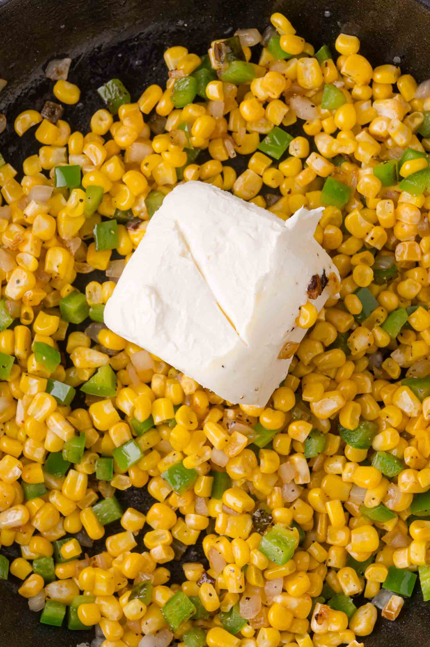 Corn kernels and chopped jalapenos in a pan with a block of cream cheese on top, ready to be mixed.
