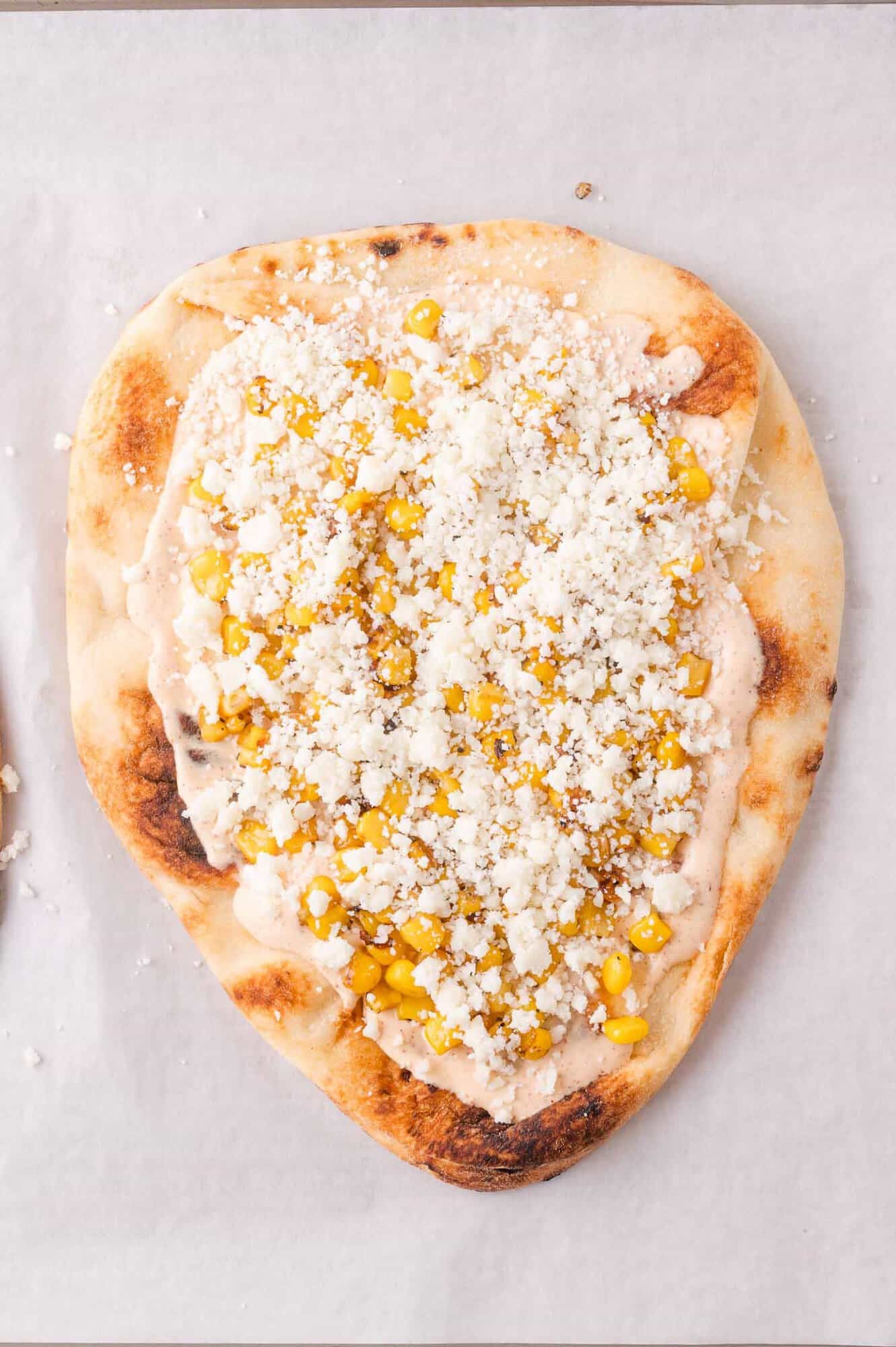 Flatbread with creamy sauce, corn kernels, and crumbled white cheese on a cookie sheet.