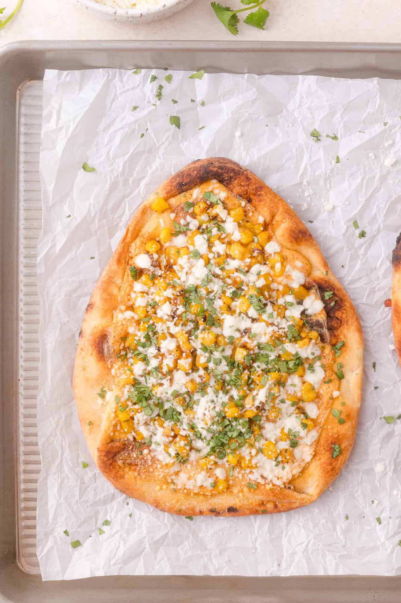 Top the elote flatbread with corn, cotija cheese, chopped cilantro, and a light sauce and place on a baking sheet lined with parchment paper.