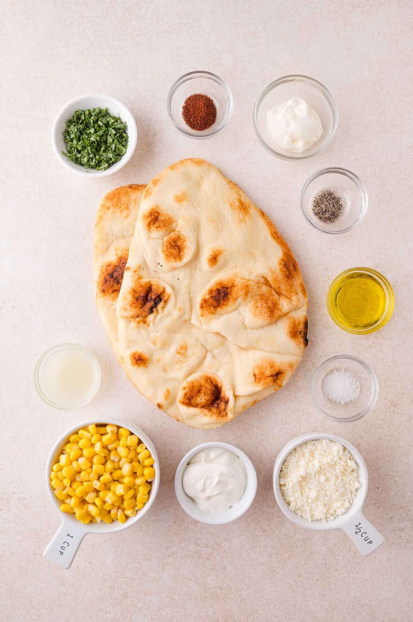 Different ingredients for flatbread dishes such as naan, corn, grated cheese, herbs, yogurt, mayonnaise, olive oil, spices, salt, pepper and lemon juice in a small bowl.