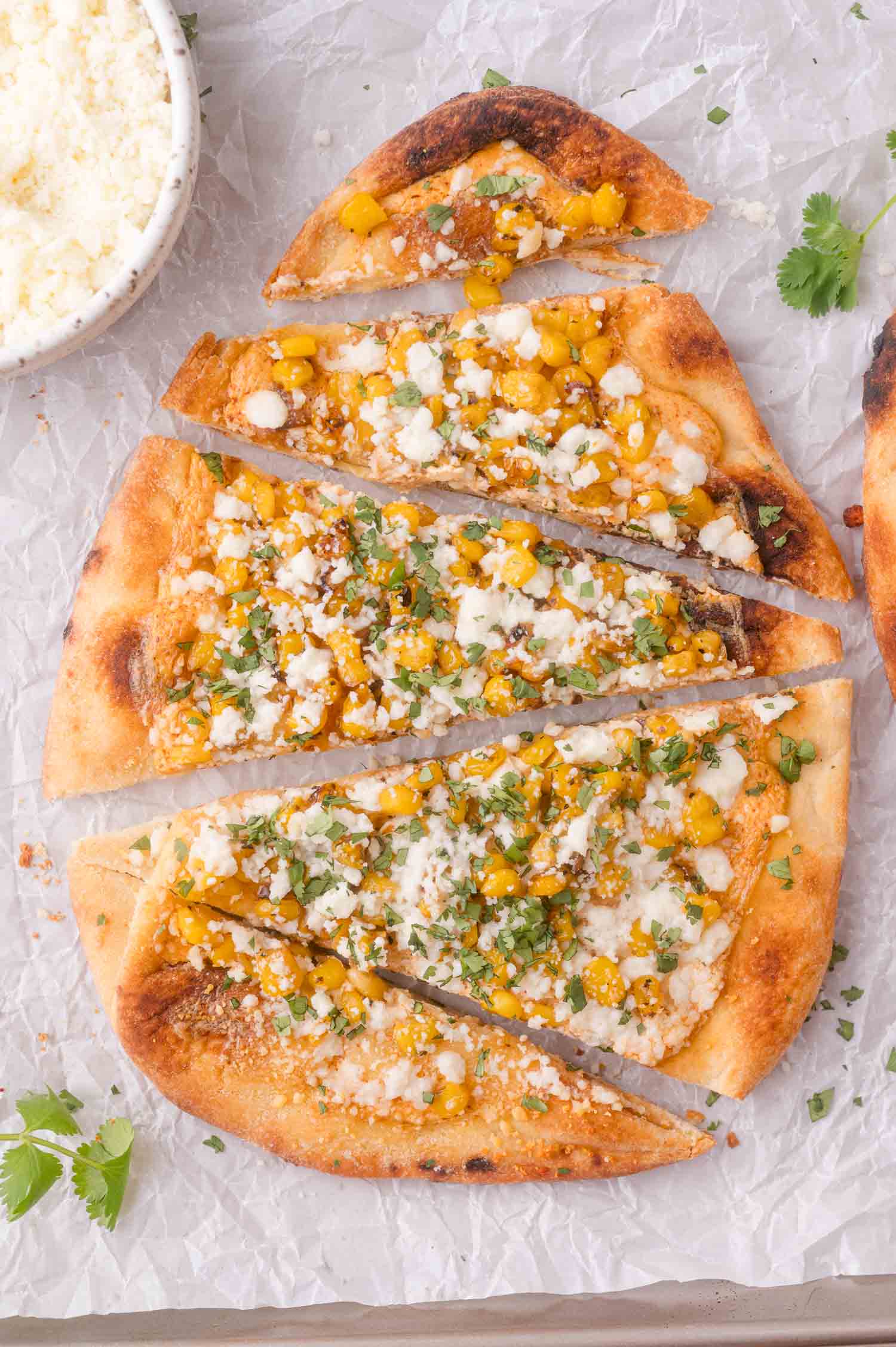 Flatbread topped with corn, cheese, herbs, and seasoning, sliced into pieces on parchment paper, with a small bowl of extra cheese nearby.