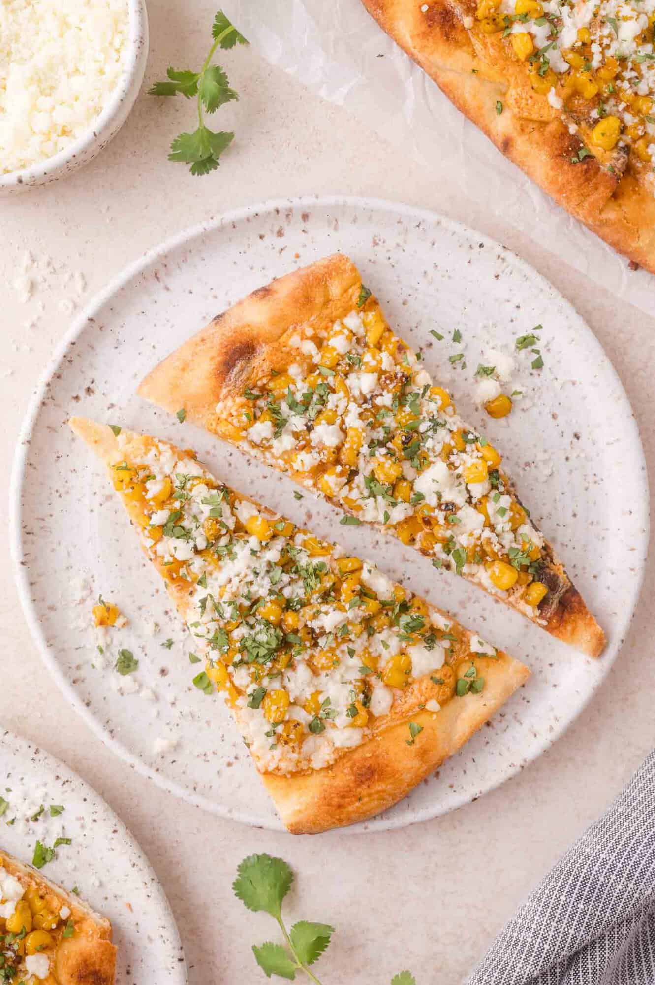 Two pieces of eloto flatbread topped with corn, crumbled cheese and chopped herbs are served on a mottled white plate.