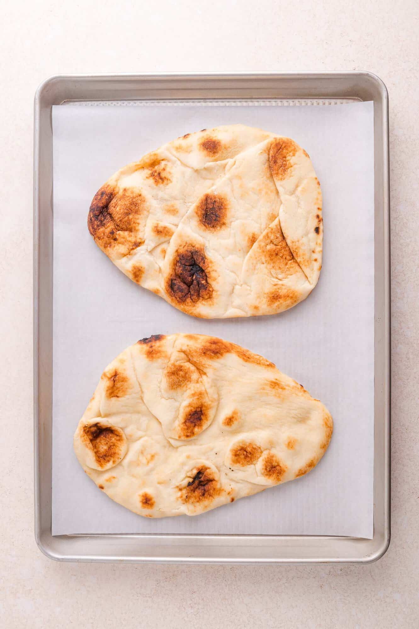 Two pieces of naan bread baked on a metal baking sheet lined with parchment paper.