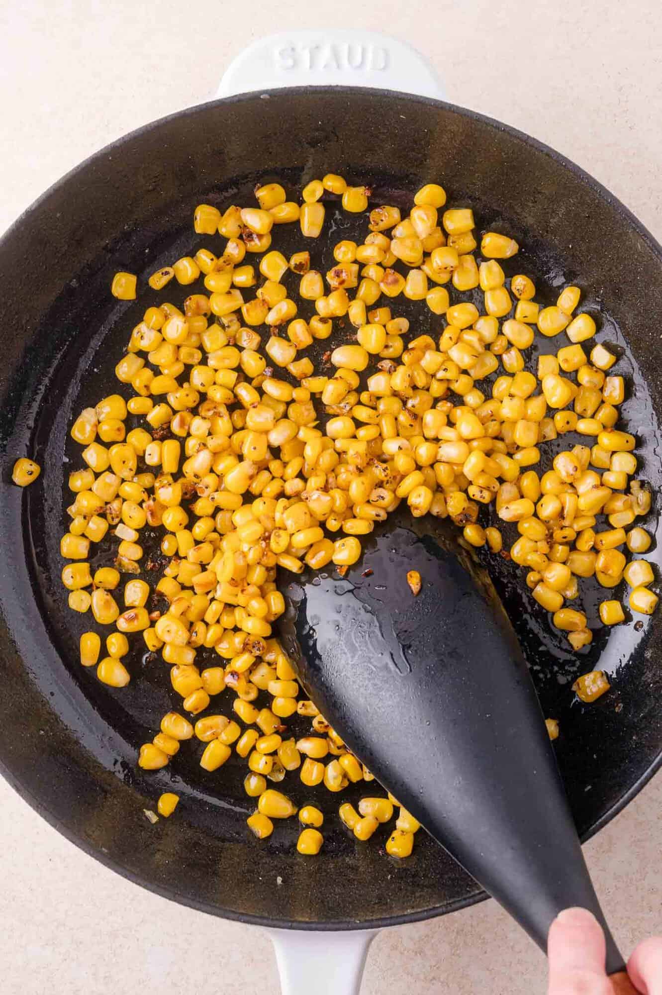 Corn kernels are stir-fried in a black frying pan with a black spatula. Some grains show browning due to cooking.