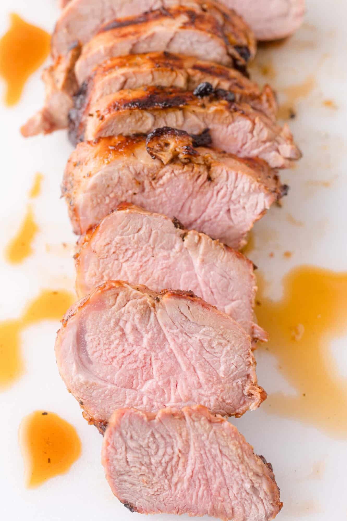 Sliced cooked pork tenderloin arranged in a row on a white surface with juices around the meat.